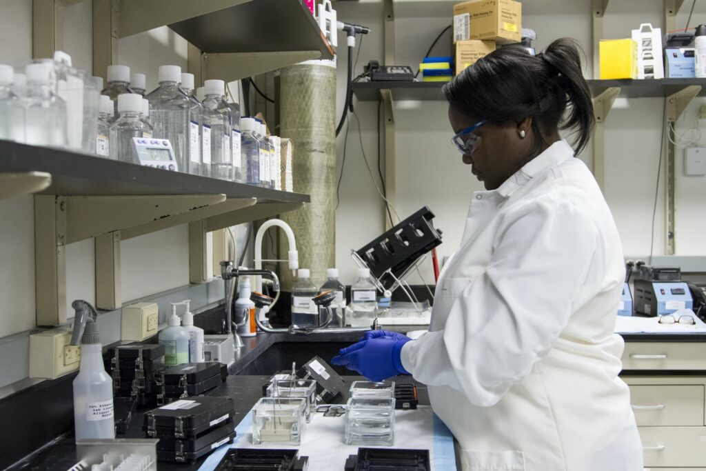 A laboratory worker in a white coat and blue gloves organizes glass containers and equipment on a lab bench